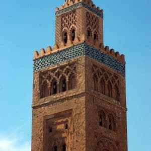 Un minarete ornamentado de piedra rojiza con tallas detalladas y azulejos geométricos azules bajo un cielo azul claro