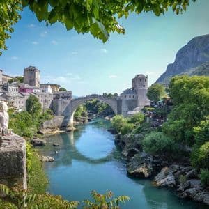 Una persona bionda siede su un muro di pietra, affacciata su un antico ponte di pietra ad arco che attraversa un fiume turchese in una città storica.