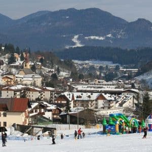 Un'ampia veduta di una località sciistica coperta di neve, con persone che sciano su una pista in primo piano e montagne boscose sullo sfondo.