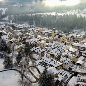 Una vista aerea di un piccolo villaggio di montagna con tetti innevati, circondato da una fitta pineta in inverno.