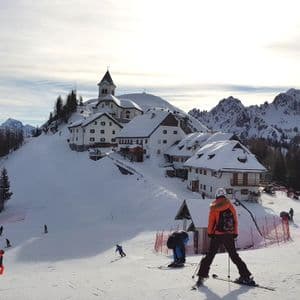 Sciatori su una pista innevata con un villaggio sormontato da una chiesa e cime montuose sullo sfondo sotto un cielo soleggiato.