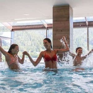 Una donna e due bambini che sorridono e schizzano acqua mentre giocano in una piscina interna di fronte a una grande finestra.