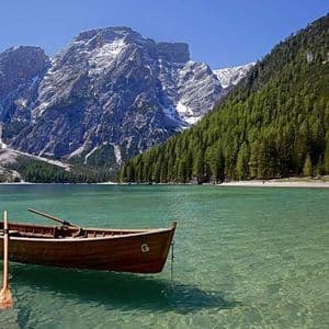Una barca a remi di legno con un remo galleggia su un lago limpido e turchese di fronte a montagne innevate e una pineta.