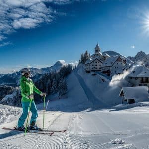 Uno sciatore con la giacca verde si erge su una pista da sci appena battuta, ammirando un paese innevato e le montagne.