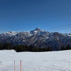 Una vista da una pista da sci innevata mostra una seggiovia che sale verso una catena di montagne innevate sotto un cielo azzurro e limpido.