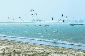 Numerosi kitesurfer planano sull'acqua increspata sotto un cielo velato, con un'ampia spiaggia sabbiosa in primo piano.