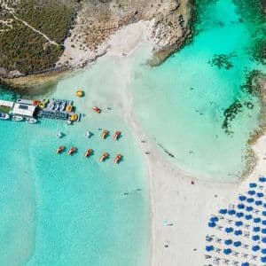 Vista aerea dall'alto di una caletta di sabbia con acqua turchese cristallina, file di ombrelloni blu e un pontile galleggiante con barche.