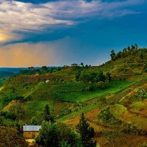 Une vaste étendue de collines verdoyantes et ondulantes, parsemée de terrasses agricoles et de quelques petites maisons, sous un ciel partagé entre soleil et nuages.