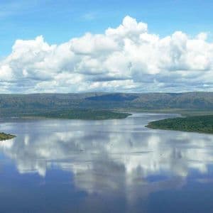 Un vaste lac calme reflète un ciel bleu avec des nuages blancs et cotonneux, entouré de vertes collines ondulantes en arrière-plan.