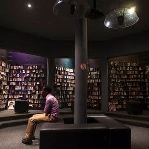 Un homme est assis sur un banc dans une salle commémorative circulaire faiblement éclairée, contemplant des murs ornés de nombreux portraits photographiques.