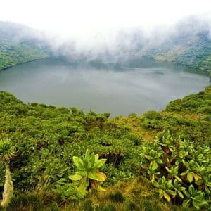 Vue plongeante d'un lac de cratère circulaire, entouré de collines à la végétation luxuriante et verte, sous un ciel brumeux et couvert.