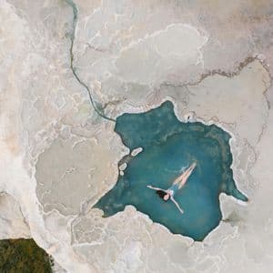 Dall'alto, una donna in costume da bagno blu galleggia sulla schiena in una piscina naturale turchese circondata da terrazze minerali bianche.