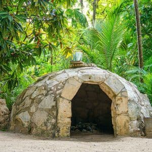 Una struttura in pietra a forma di cupola con un ingresso ad arco si trova su un terreno sabbioso, circondata da una lussureggiante vegetazione tropicale.