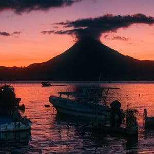 Diverse barche galleggiano su un lago al tramonto con una grande montagna in silhouette sullo sfondo sotto un cielo arancione e viola.