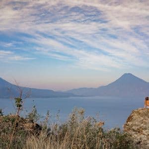 Una persona con un cappello è seduta su una grande roccia, ammirando un vasto lago circondato da montagne sotto un cielo nuvoloso.