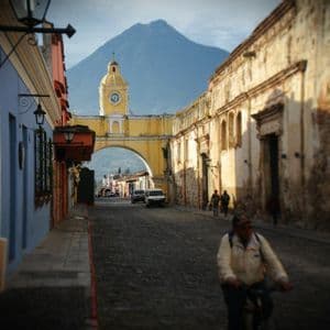 Una persona in bicicletta percorre una strada acciottolata verso un arco giallo con una torre dell'orologio, con un grande vulcano sullo sfondo.