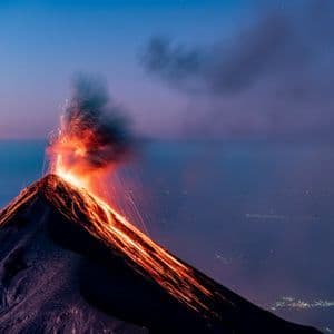 Un vulcano erutta al tramonto, con lava fusa che scorre lungo i suoi pendii e scintille che si alzano nel cielo sopra un paesaggio di luci cittadine.