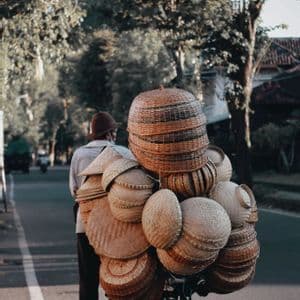 Una persona, ripresa di spalle, spinge una bicicletta carica di cesti intrecciati lungo una strada alberata.