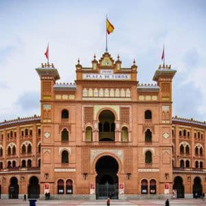 La facciata simmetrica in mattoni rossi dell'edificio della Plaza de Toros, con archi e una bandiera spagnola, vista da una grande piazza sotto un cielo nuvoloso.