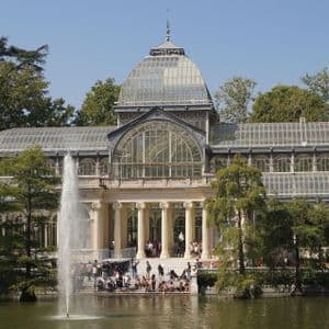 Un sontuoso palazzo di vetro con cupola sorge in riva a un lago, con un'alta fontana che zampilla acqua in primo piano.