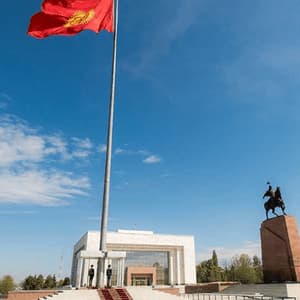 La bandiera del Kirghizistan sventola su un'alta asta in una piazza, con due guardie alla sua base davanti a un edificio moderno e a una statua equestre.