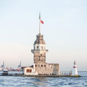 Una torre di pietra con una bandiera turca si erge su una piccola isola nel mare, con una città portuale visibile in lontananza.