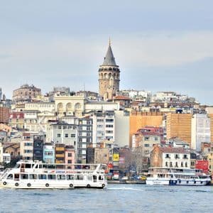 Un traghetto bianco naviga sull'acqua con la Torre di Galata e un denso skyline cittadino sullo sfondo sotto un cielo pallido.