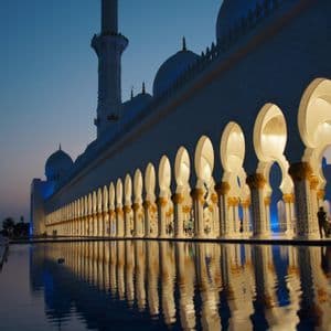 Una moschea bianca illuminata con un colonnato di archi e un minareto che si riflette nell'acqua calma al crepuscolo.