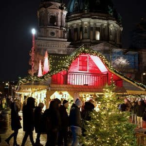 Menschen gehen nachts über einen Weihnachtsmarkt, vorbei an beleuchteten Ständen und einem beleuchteten Baum, mit einem großen Kuppelgebäude im Hintergrund.
