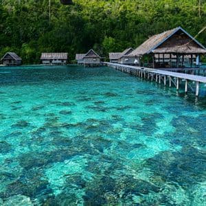 Un lungo pontile in legno collega bungalow sull'acqua sopra un mare turchese cristallino, con un'isola verde e lussureggiante sullo sfondo.