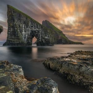 Scatto a lunga esposizione di due grandi faraglioni, uno con un arco naturale, su una costa rocciosa sotto un drammatico cielo al tramonto.