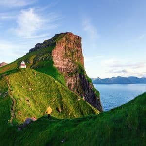 Una persona in giacca gialla cammina su un sentiero lungo una verde scogliera costiera con un faro in lontananza.