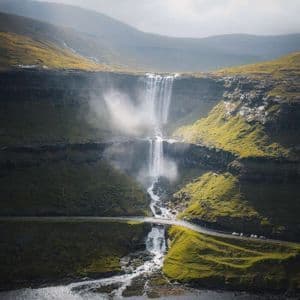 Una cascata a più livelli scende da scogliere verdi, scorre sotto una strada con auto e si getta in uno specchio d'acqua sottostante.