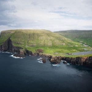 Una vista aerea di una grande scogliera verdeggiante su una costa frastagliata, con un piccolo villaggio e un lago sullo sfondo sotto un cielo nuvoloso.