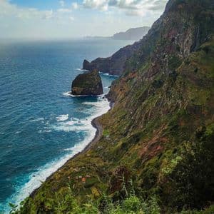 Una ripida scogliera verde incontra l'oceano blu lungo una costa frastagliata, con onde che lambiscono una piccola spiaggia e un villaggio sottostante.