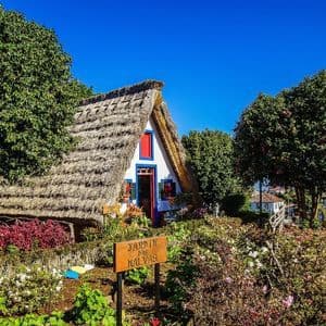 Una casa tradizionale a forma di A con tetto di paglia si trova in un rigoglioso giardino con fiori e alberi colorati sotto un cielo azzurro e limpido.