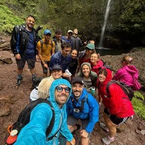 Un gruppo WeRoad in impermeabili colorati posa per un selfie davanti a un'alta cascata in una foresta lussureggiante.
