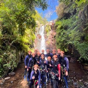 Un gruppo WeRoad in viaggio, con attrezzatura completa da canyoning, in posa in un torrente roccioso alla base di una cascata.