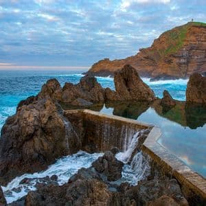 Una piscina naturale incastonata tra rocce vulcaniche sulla costa, con l'acqua che tracima nell'oceano mentre le onde si infrangono lì vicino.