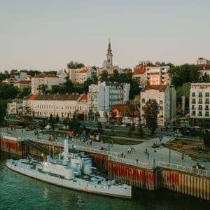 Una nave bianca è ormeggiata a un molo cittadino lungo un fiume, con un paesaggio urbano storico e un campanile di chiesa sullo sfondo al tramonto.