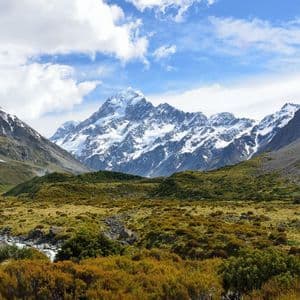 Un'imponente catena montuosa innevata domina un'ampia valle, ricca di arbusti color oro e un ruscello roccioso, sotto un cielo blu con nuvole soffici.