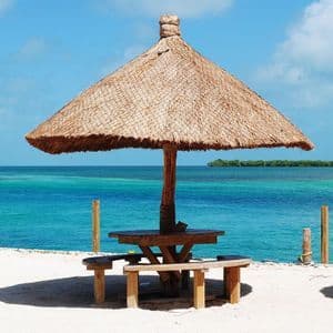 Un tavolo da picnic in legno con panche si trova sotto un grande ombrellone di paglia, su una spiaggia di sabbia bianca con vista su un oceano turchese.