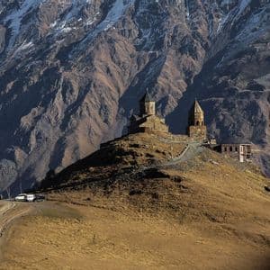 Una chiesa e un campanile in pietra si ergono su una collina erbosa, con una massiccia e aspra catena montuosa sullo sfondo.
