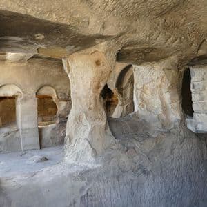 L'interno di un'abitazione rupestre scavata a mano con pilastri in pietra e finestre ad arco sotto un soffitto di roccia grezza.
