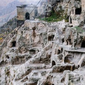 Un monastero rupestre a più livelli scavato in una parete rocciosa, con scalinate e passerelle che si affacciano su una valle montuosa.