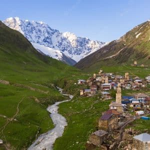 Una vista aerea di un piccolo villaggio con torri in pietra in una valle verde, situato vicino a un fiume con montagne innevate sullo sfondo.