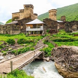 Un ponte di legno attraversa un fiume impetuoso che conduce a un antico villaggio con torri di pietra su un pendio verdeggiante ed erboso.