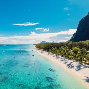 Vista aerea di una costa tropicale con spiaggia di sabbia bianca, palme, acqua turchese cristallina e una montagna verde.