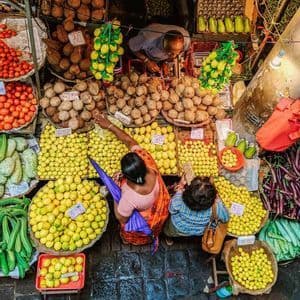 Vista dall'alto di due donne che acquistano limoni e noci di cocco da un venditore in un affollato mercato all'aperto.