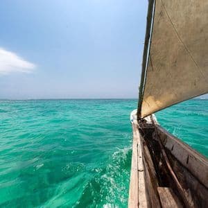 Vista dalla prua di una barca a vela in legno con vela di tela, che naviga su acque turchesi sotto un cielo azzurro.